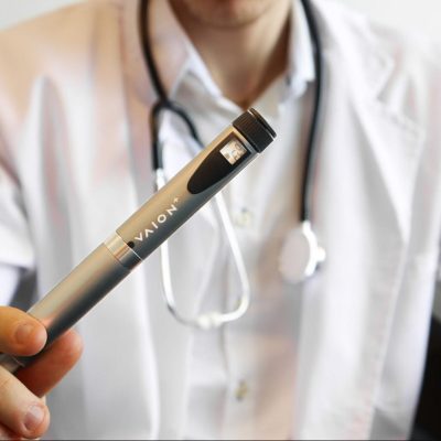 A person wearing a white lab coat and a stethoscope is holding a VAION pen. The pen is metallic with the brand name "VAION+" visible on its body.